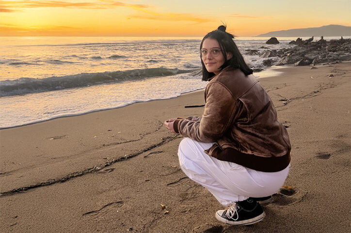 A young woman squats on the shoreline of a beach at sunset.