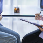 A teen sits across from a doctor. The teen holds their stomach in discomfort while the doctor takes notes.