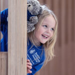 A happy, smiling five-year-old girl, holding her teddy bear, peeks playfully around a corner.