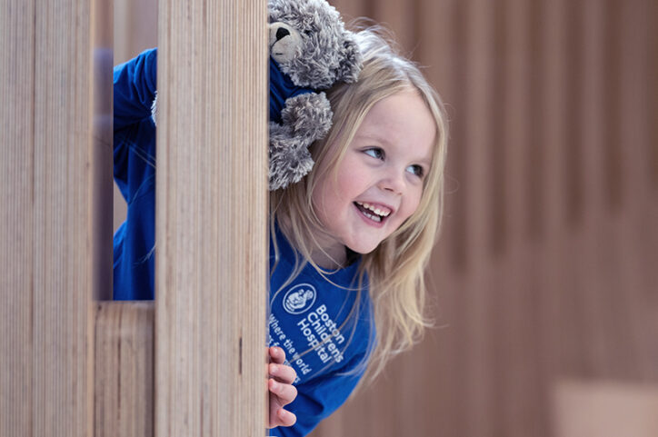 A happy, smiling five-year-old girl, holding her teddy bear, peeks playfully around a corner.