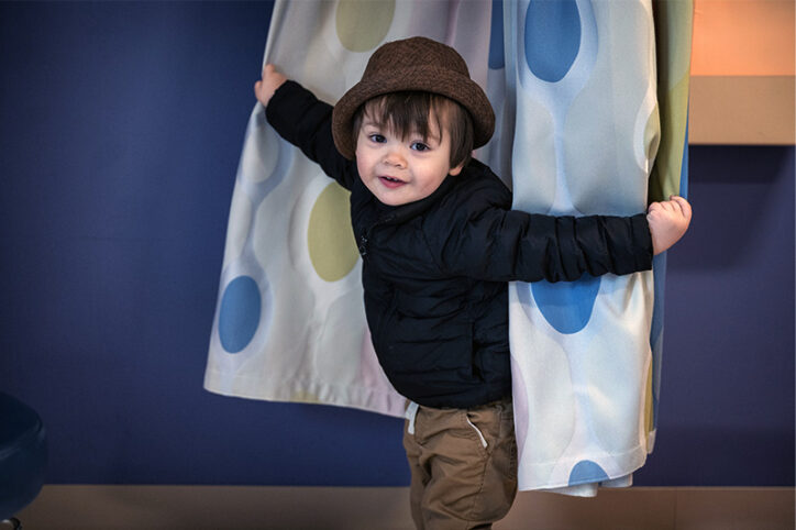 2-year-old boy who had brachial plexus birth injury peers out of a curtain in a medical exam room.