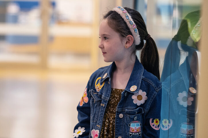 A 6-year-old wearing a jean jacket stands in a hospital hallway, gazing into the distance