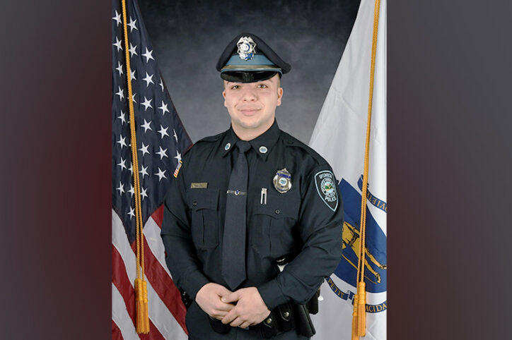 Jack stands in his police uniform before an American flag, on left, and Massachusetts state flag, on right.