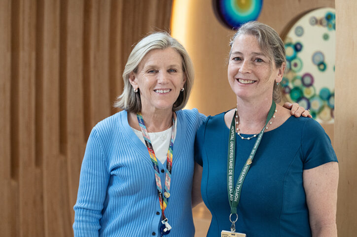 Two nurses smiling and standing side by side, wearing ID badges and lanyards, one with her arm around the other.