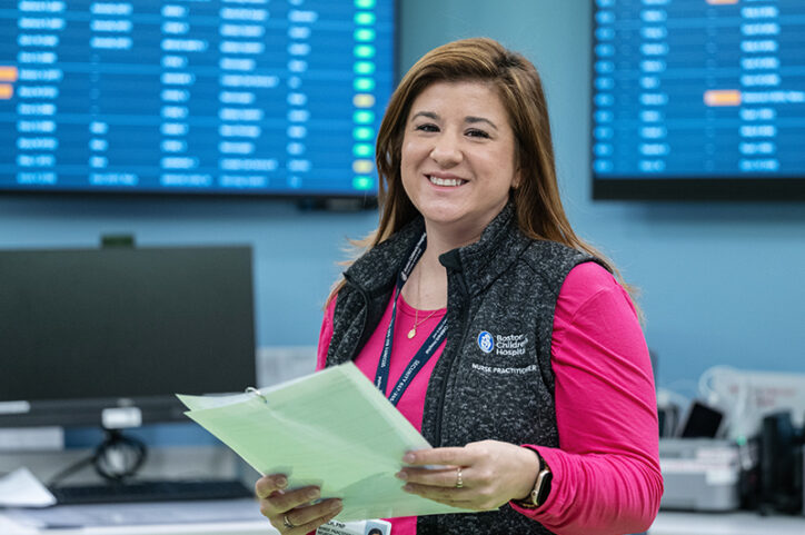 A nurse practitioner smiles while holding papers in a hospital unit, with monitors and computers behind her.