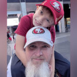 A photo of a little boy sitting on his father's shoulders. Both are wearing hats with the San Francisco 49ers logo.