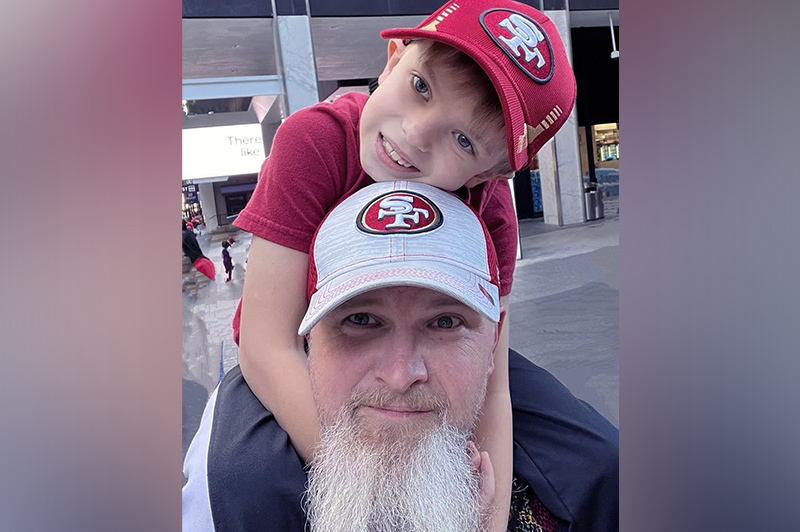 A photo of a little boy sitting on his father's shoulders. Both are wearing hats with the San Francisco 49ers logo.