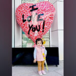 A 2-year-old girl stands in front of a large heart-shaped sculpture that reads “I LOVE YOU!”