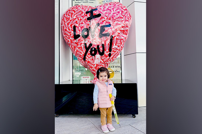A 2-year-old girl stands in front of a large heart-shaped sculpture that reads “I LOVE YOU!”
