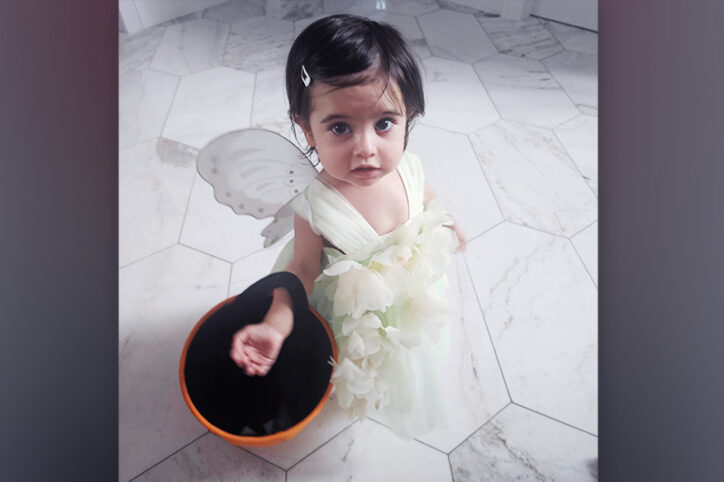 A 2-year-old girl in a pale green flower dress and butterfly wings holds a Halloween bucket.