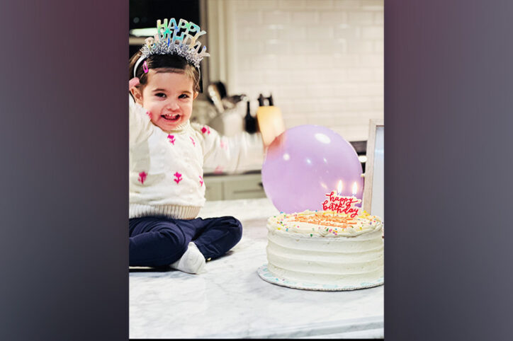Two-year-old girl wearing a birthday crown sits beside a cake and a purple balloon on a kitchen counter.