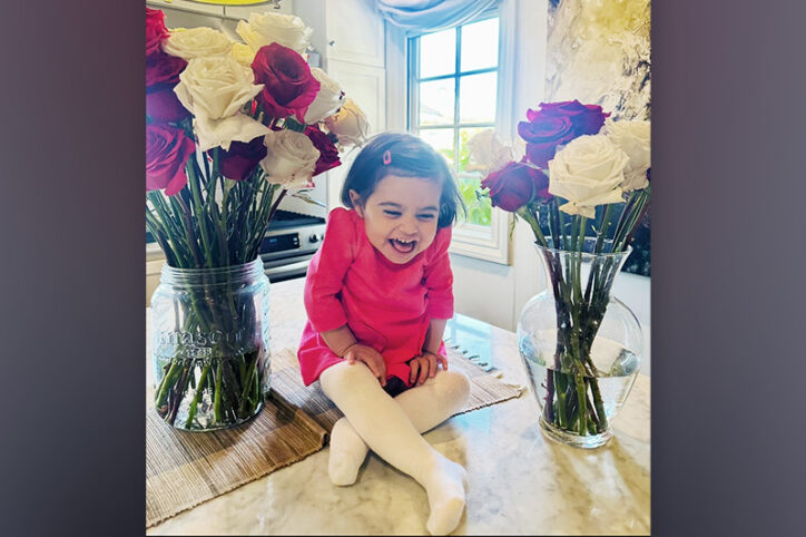 A 2-year-old girl wearing a pink dress and white tights sits on a kitchen counter, laughing.