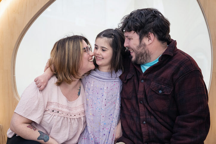 A dark-haired 5-year-old girl sits between her mother and father, smiling