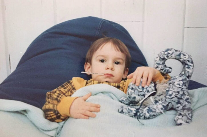 A young Stephan pats the head of a stuffed animal while laying in bed and having his temperature taken orally.
