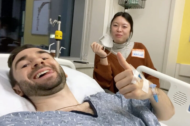 Stephan gives a thumbs-up while lying in a hospital bed. His wife sits next to the bed.