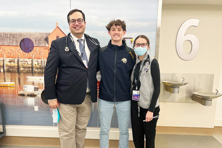 A teenage boy poses smiling with two doctors in a hospital hallway.