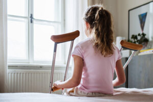 A sad-looking girl with crutches looks out a bedroom window.