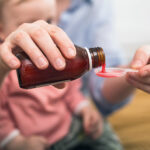 A person pouring medicine into a spoon while a child sits on their lap.