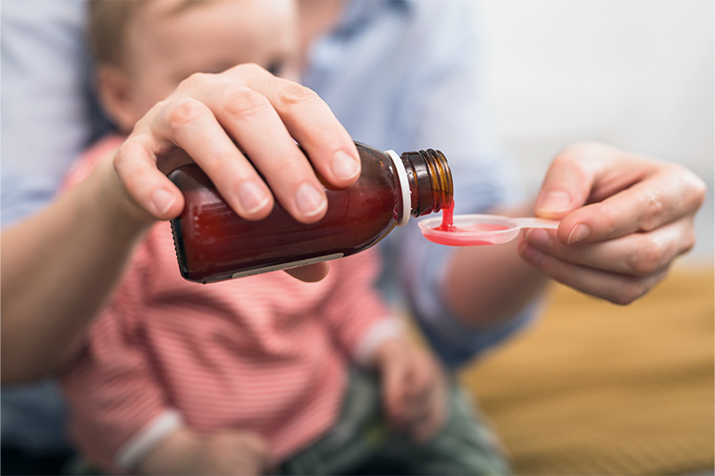 A person pouring medicine into a spoon while a child sits on their lap.