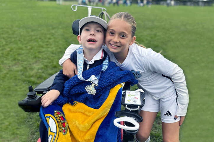 A school-age girl in a soccer uniform puts her arm around her brother in a wheelchair as both smile.