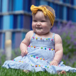 A baby girl with a bow on her head, sitting in the grass with a blue building in the background.