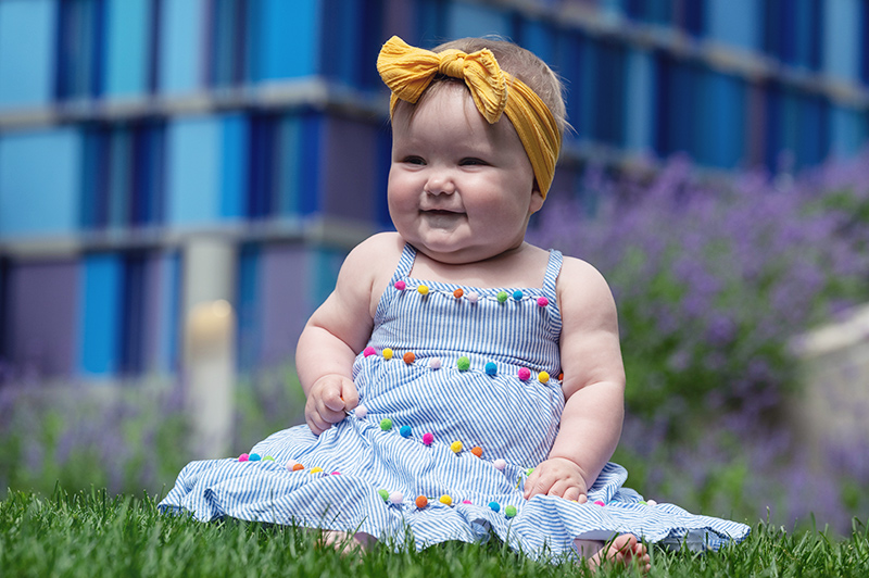 A baby girl with a bow on her head, sitting in the grass with a blue building in the background.