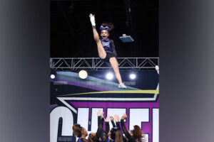 A cheerleader is tossed into the air with one leg extended and arms raised.