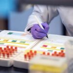 a hand in a latex glove taking notes near a batch of test tubes