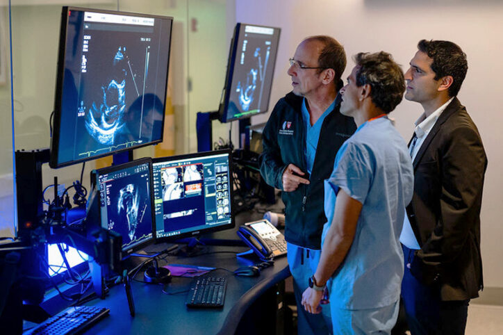 Three cardiologists stand while looking at a large-screen computer image of an echocardiogram.