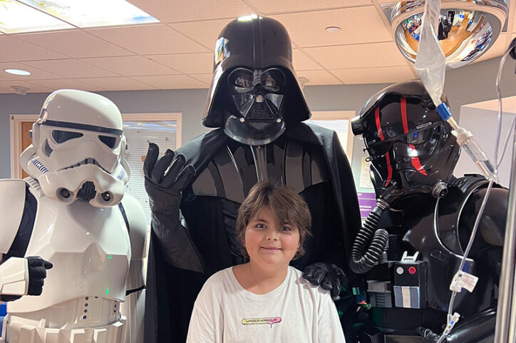 A preteen boy standing with people dressed as Darth Vader and stormtroopers.