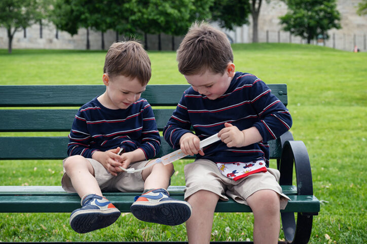Two young boys sit on a park bench while one helps the other receive formula through a feeding tube