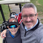 A preteen boy stands with his father in front of a golf cart
