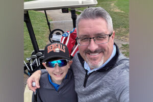 A preteen boy stands with his father in front of a golf cart