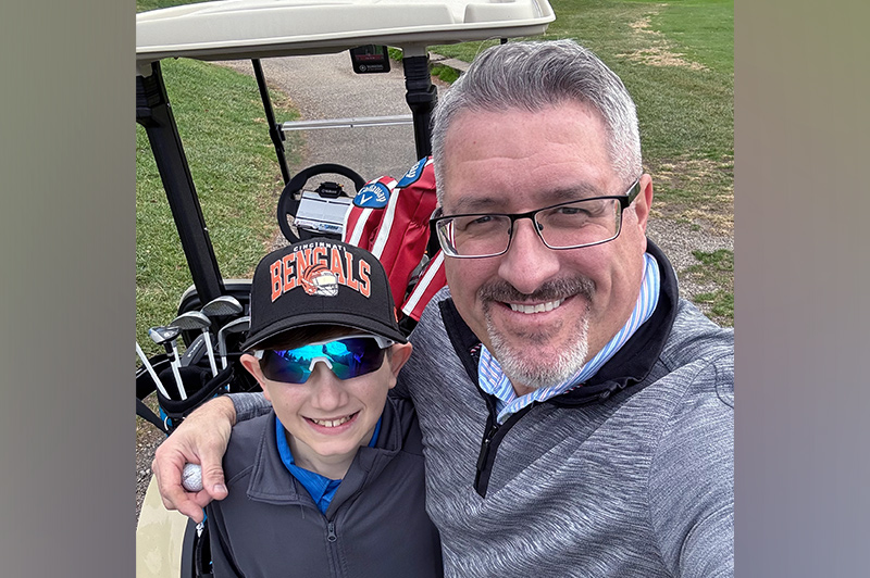 A preteen boy stands with his father in front of a golf cart