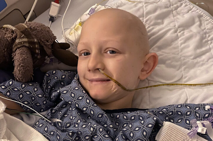 A bald preteen boy in a hospital bed with a feeding tube in his nose