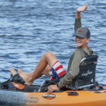 A 12-year old boy smiles and waves from a pedal kayak.
