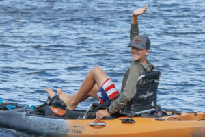 A 12-year old boy smiles and waves from a pedal kayak.