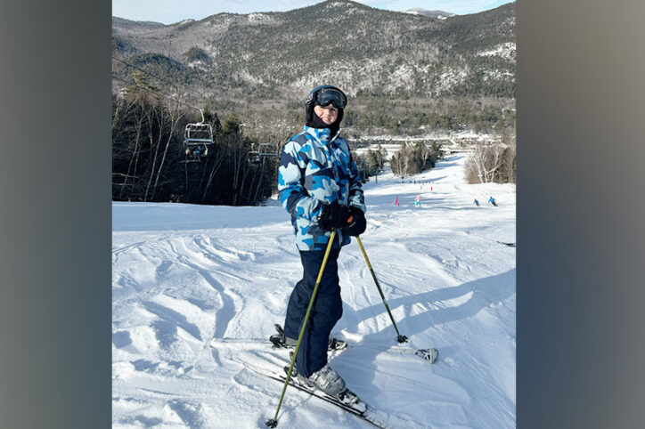 A smiling 11-year-old boy on skis at the top of a ski slope.