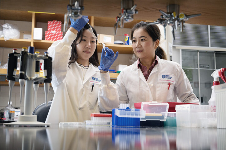 One female researcher in a lab coat carefully transfers a sample into a vial while another observes.