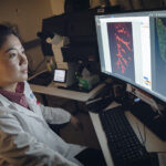 A female researcher in a lab coat examines images of tissue samples on a computer screen.