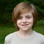 Young boy with shaggy brown hair smiling wearing a light gray shirt and standing in front of a blurred green background.