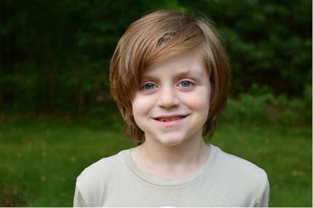 Young boy with shaggy brown hair smiling wearing a light gray shirt and standing in front of a blurred green background.