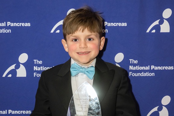 A young boy in a tuxedo holds an award, standing in front of a blue backdrop with the National Pancreas Foundation logo.