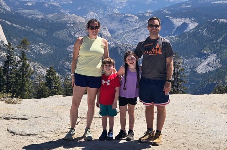 A family of four stands on a rocky ledge with a mountain view in the background. 