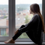 A woman sits on a windowsill, gazing outside at blurred buildings.