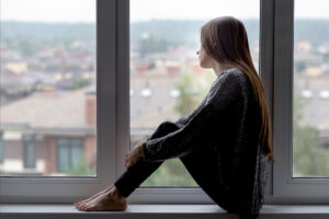 A woman sits on a windowsill, gazing outside at blurred buildings.