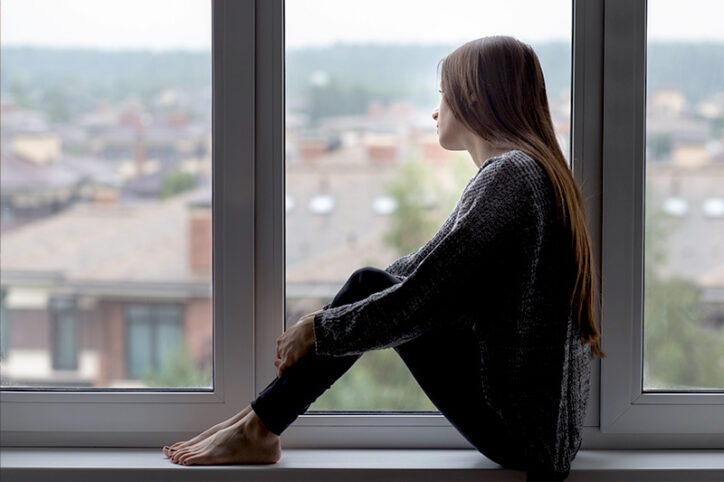 A woman sits on a windowsill, gazing outside at blurred buildings.