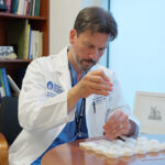 A man in a white lab coat sitting at a table dropping liquid into a tray case.