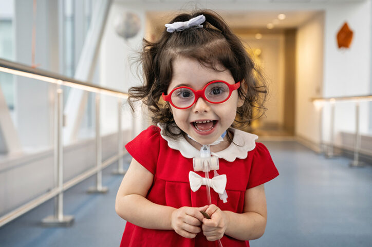 A young girl with a tracheostomy smiles while standing in a hospital hallway.
