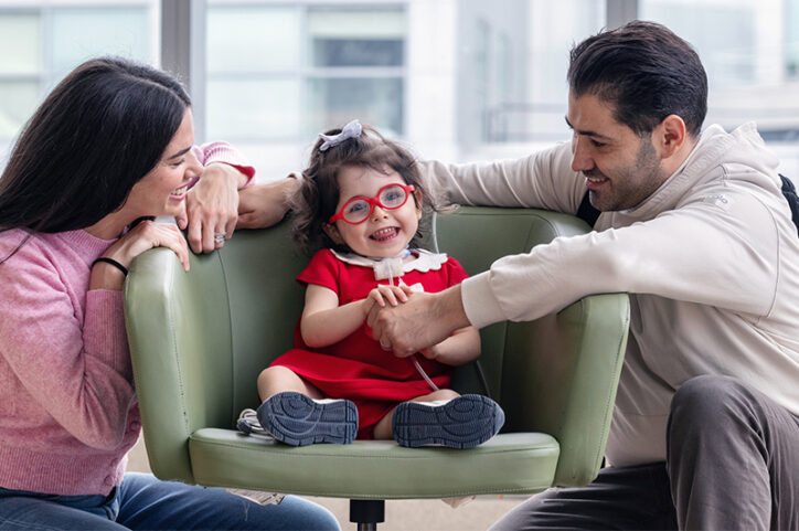 A young girl with a tracheostomy sits on a chair between two adults, smiling.